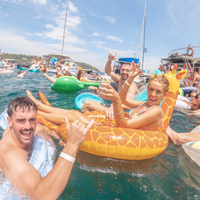 A group of people enjoy a party on the water, relaxing on colorful inflatables including a giraffe float. They smile and make hand signs, with boats and blue sky in the background.
