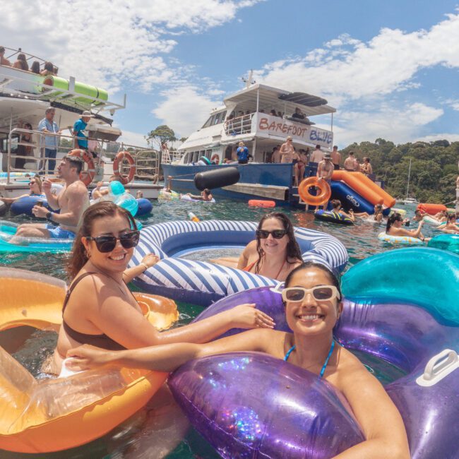 A group of smiling people relax on colorful inflatable floaties in the water near boats during a lively outdoor party under a partly cloudy sky.