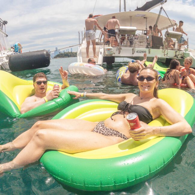 People relaxing on colorful inflatable loungers in the water near a yacht, holding drinks and smiling. Several others are gathered on the yacht and in the water, enjoying a sunny day.
