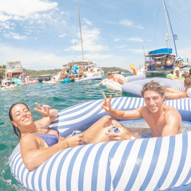 Two people in swimsuits smile and flash peace signs while lounging in a striped inflatable on blue water, surrounded by others floating and boats on a sunny day.