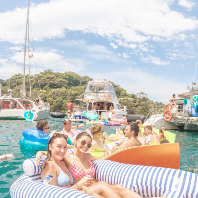Two women in swimsuits smile while relaxing on a striped inflatable float in the water. They are surrounded by others on colorful floats and boats, with a tree-covered island and blue sky in the background.