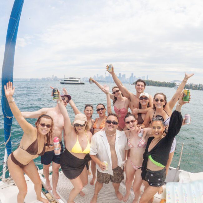 A group of people in swimsuits stand on a boat deck, smiling and raising drinks. The city skyline and water are visible in the background under a partly cloudy sky.