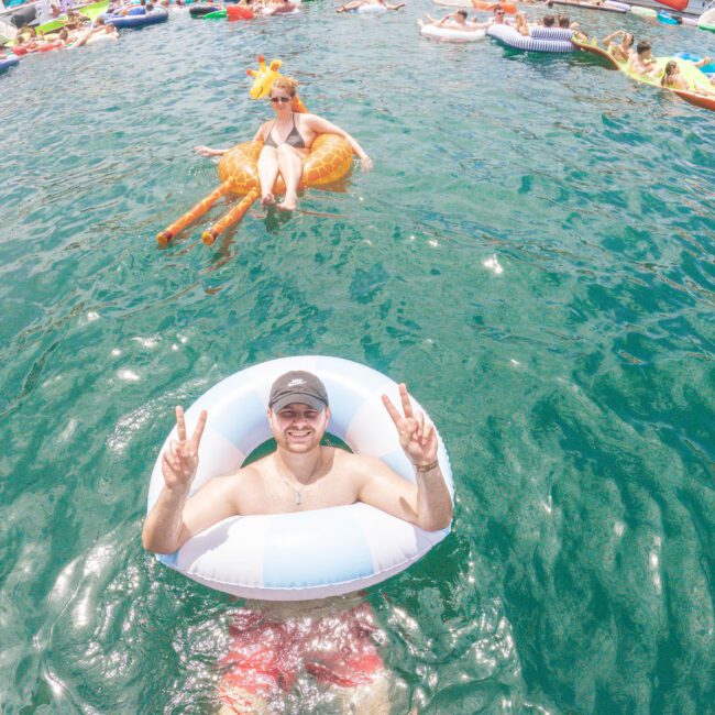 A man in a white and blue float tube flashes peace signs in clear blue water, while people relax on various inflatables and boats in the background during a lively summer party.