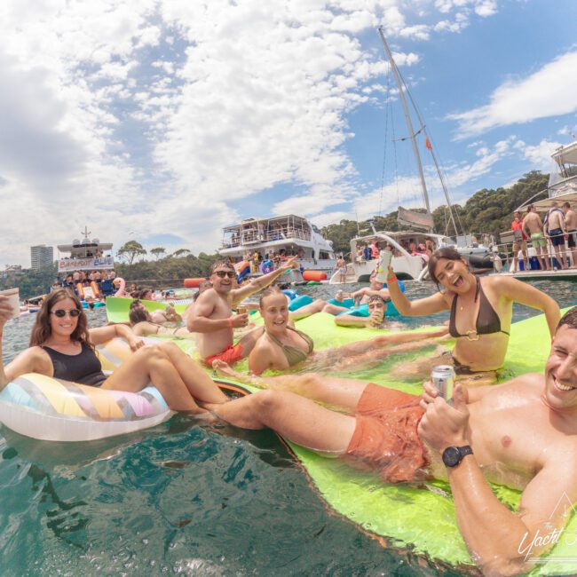 A group of smiling people relax on colorful floaties in the water near boats, holding drinks and enjoying a sunny day. Trees and blue sky with clouds are visible in the background.