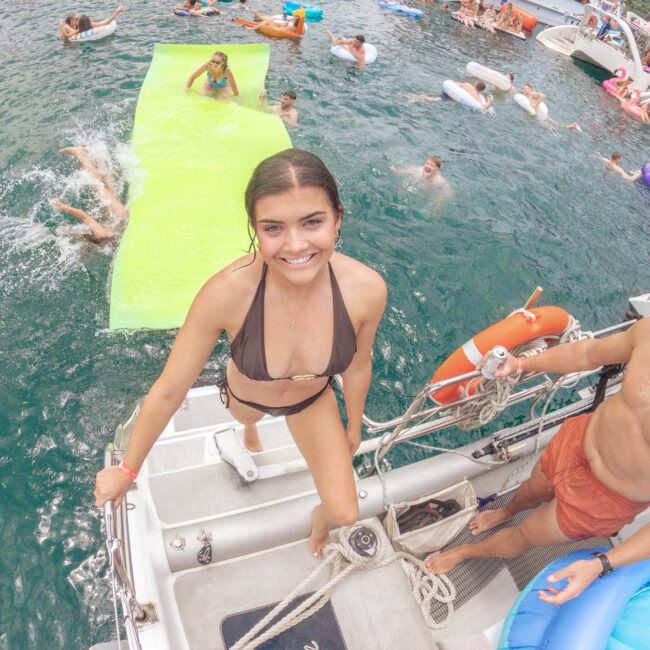 A smiling woman in a bikini stands on a boat ladder, with people swimming and lounging on floaties and rafts in the water. Several boats and a lively party atmosphere are visible in the background.
