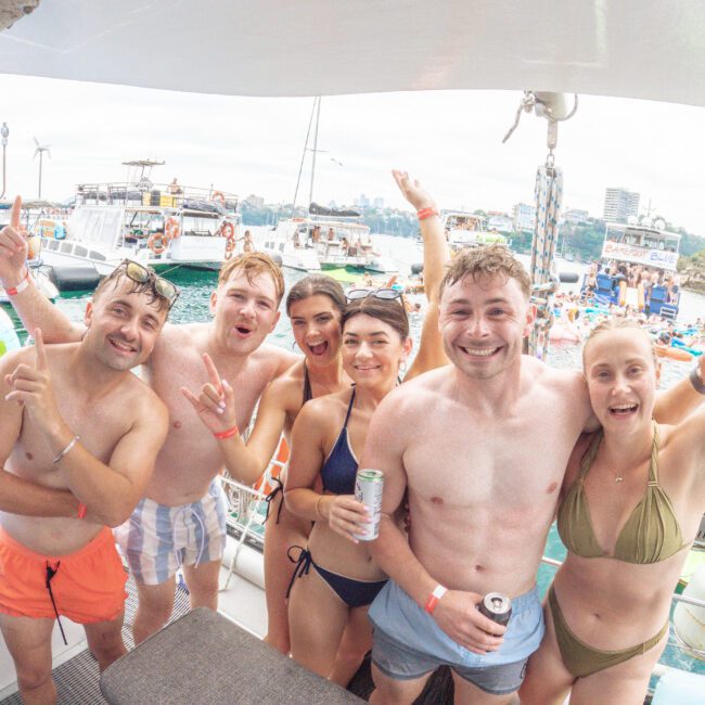 Six young adults in swimwear smile and pose for a group photo on a boat, holding drinks. Other boats and people are visible in the background, suggesting a lively party atmosphere on the water.