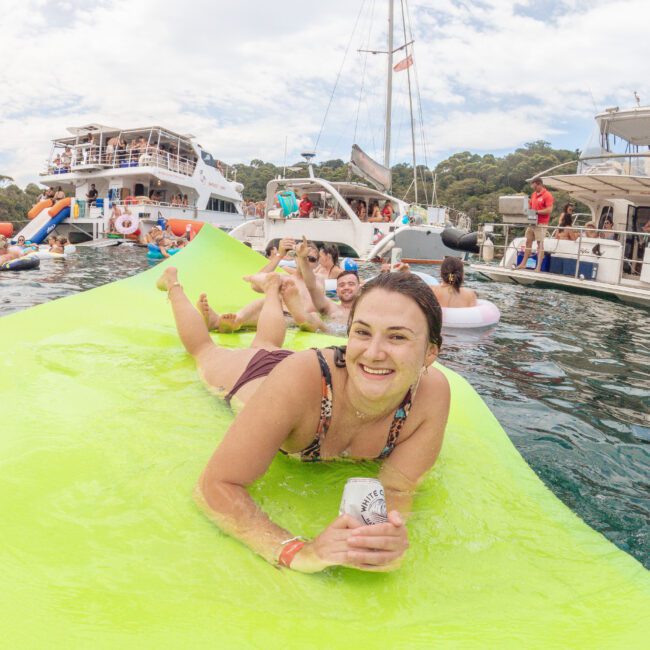 A smiling woman lounges on a bright green inflatable mat in the water, holding a drink. Other people relax nearby on the mat and in pool floats, with boats and yachts anchored in the background. The atmosphere is festive.