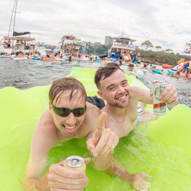 Two men in sunglasses relax on a bright green float in the water, smiling and holding cans at a lively boat party. Many boats and people can be seen celebrating in the background under a cloudy sky.