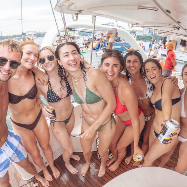 A group of smiling young adults in swimsuits pose together on a boat, holding drinks. Water, other boats, and a city skyline are visible in the background, suggesting a lively summer outing.