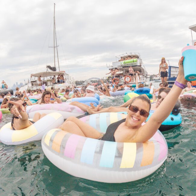 A crowd of people relaxing on colorful inflatable tubes in the water at a lively boat party, with boats and cloudy skies in the background. A smiling woman in sunglasses raises a drink in the foreground.
