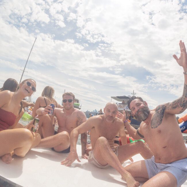 Four adults in swimsuits sit on a floating platform, smiling and waving at the camera. Boats and other people are visible in the background under a partly cloudy sky. The atmosphere is lively and festive.