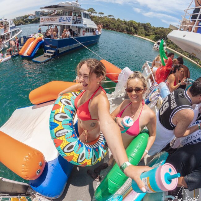 Two women in swimsuits with pool floats and drinks smile at the camera on a boat during a lively summer party; other people and boats are seen around them on the water in bright, sunny weather.