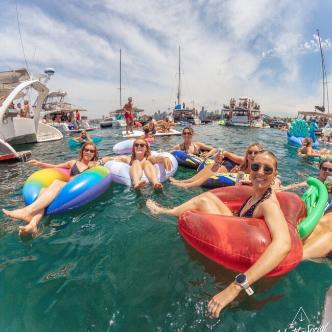 A group of women in sunglasses relax on colorful pool floats in the water near boats and yachts, smiling at the camera under a sunny, blue sky.