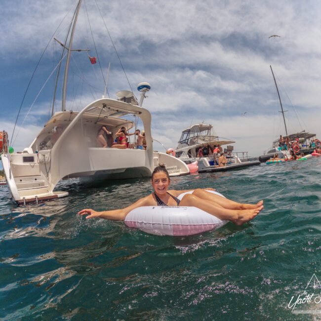 A woman smiles while floating on an inflatable ring in the water near several anchored boats filled with people enjoying a sunny day. The sky is clear and the atmosphere is lively and festive.