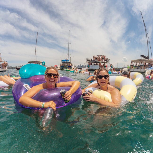 Two women in sunglasses smile while relaxing on colorful inflatable floats in the water, holding drinks. Boats with people are visible in the background under a partly cloudy sky. The Yacht Social Club logo appears in the corner.