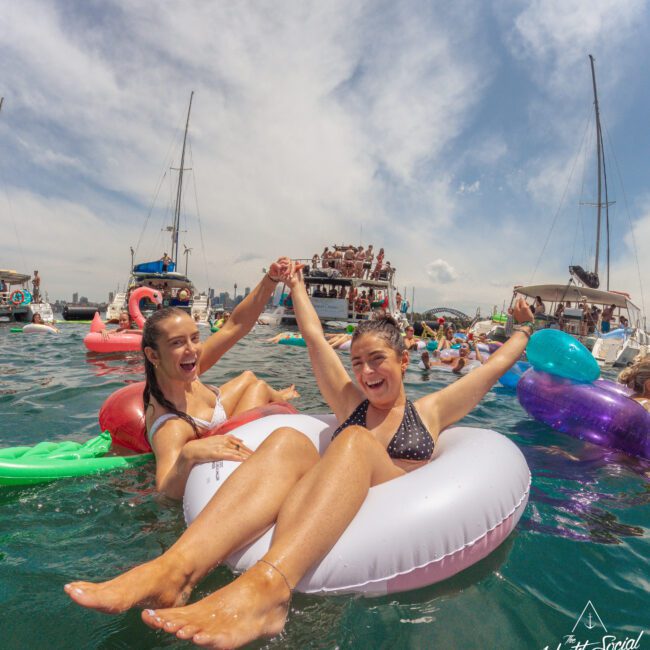 Two smiling women relax on colorful inflatable floats in the water, holding hands with arms raised. Other people and boats are visible in the background during what looks like a lively outdoor summer event.