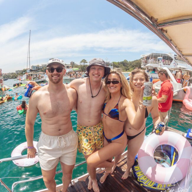 Four smiling people in swimsuits stand on a boat deck, holding drinks, with water, other boats, and people partying in the background. It’s sunny and festive, with pool floats and drinks visible.