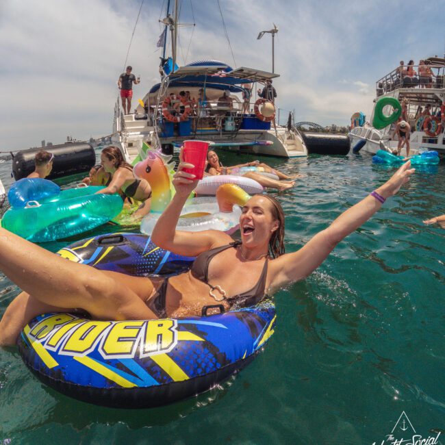 A woman in a bikini smiles and raises her arm while floating on an inflatable tube in the water near a yacht party with people socializing and relaxing on floats under a sunny sky.