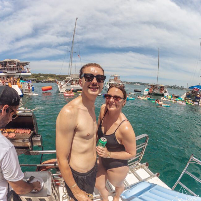 A smiling man and woman in swimsuits stand on a boat deck holding drinks, with other people, boats, and inflatables on the water behind them under a partly cloudy sky.