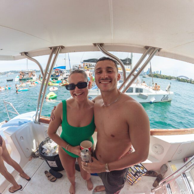 A smiling woman in a green swimsuit and a shirtless man stand together on a boat, holding drinks. Behind them, people float and relax on boats in turquoise water under a sunny sky.