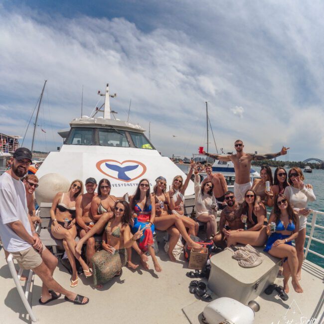 A group of people in swimwear pose and smile together on the deck of a yacht, with boats and a marina visible in the background under a partly cloudy sky.
