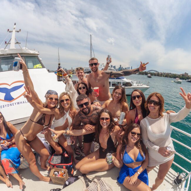 A group of friends in swimsuits and summer clothes smile and pose with drinks on a boat under sunny skies, with water and a city skyline in the background.