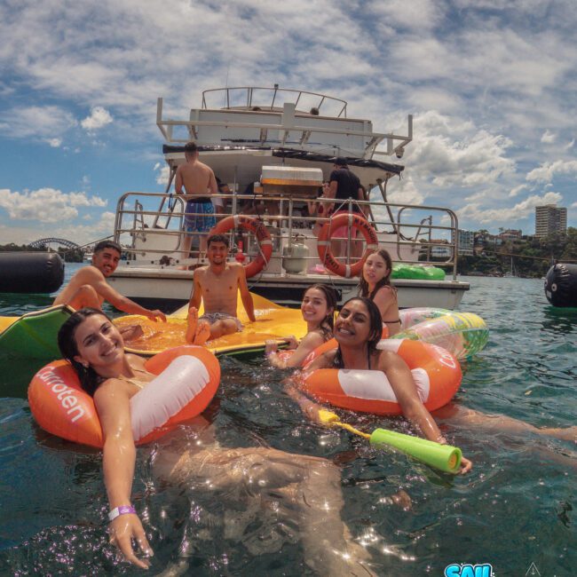 A group of young people in colorful pool floats relax and smile in the water near a boat on a sunny day, with more people visible on the boat and city buildings in the background.