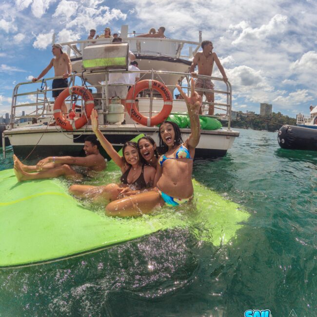 Three women in swimsuits smile and pose on a floating mat in the water near a boat, with others on the boat enjoying the sunny weather. The sky is partly cloudy, and buildings are visible in the background.