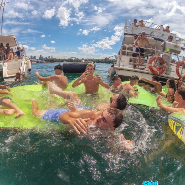 A group of kids play and swim on a bright green floating mat in the water, surrounded by boats under a blue sky with scattered clouds. Some children are splashing, while others watch from the boats.