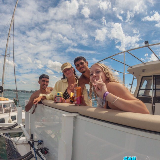 Four young adults smiling and holding drinks while leaning on the railing of a boat under a partly cloudy sky. The ocean and another boat are visible in the background. "Sail Gustavo" logo appears in the bottom right corner.
