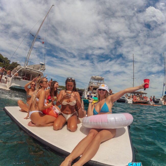 A group of women in swimsuits relax on a floating mat in the sea near boats, enjoying drinks and sunshine. One woman wears a float and raises her cup, smiling. The sky is partly cloudy. The logo "Sail Gustavo" is visible.