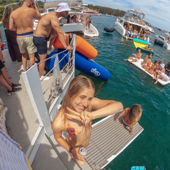 A young woman in a bikini flashes a peace sign and smiles on a boat deck during a lively boat party, with people enjoying the water, boats, and inflatables in the background. The Sail Gustavo logo is visible in the corner.