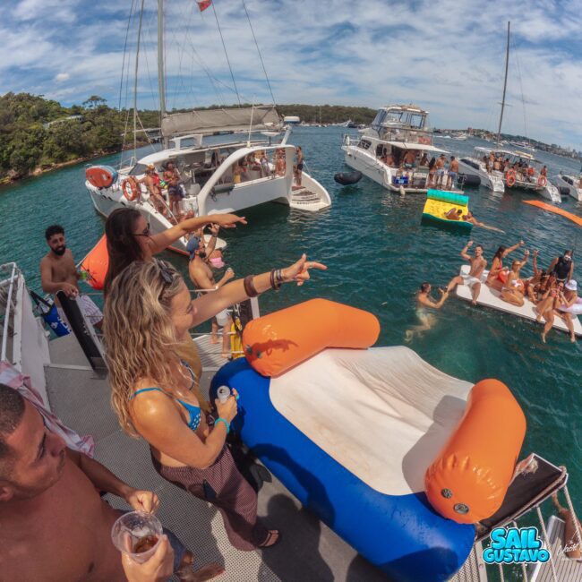 People on boats and a floating inflatable slide enjoy a lively party on the water, with groups swimming, relaxing, and socializing under sunny skies. The scene is vibrant and festive.