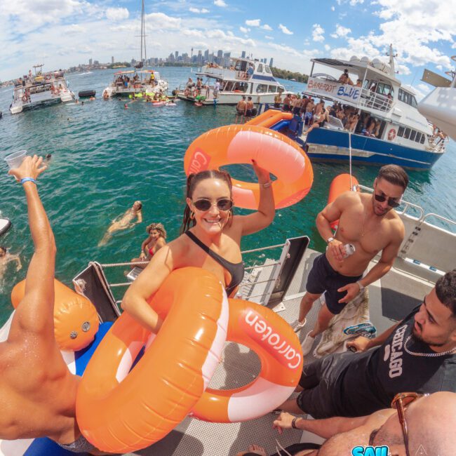 A woman wearing sunglasses and an inflatable ring smiles on a boat with other people during a lively daytime pool party on the water; boats and a city skyline are visible in the background.