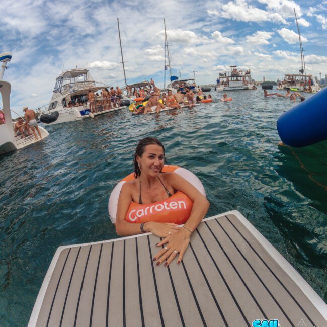 A woman in an orange Carroten float relaxes by a floating dock in the ocean, smiling at the camera. Several boats and people enjoying the water are visible in the background under a partly cloudy sky.