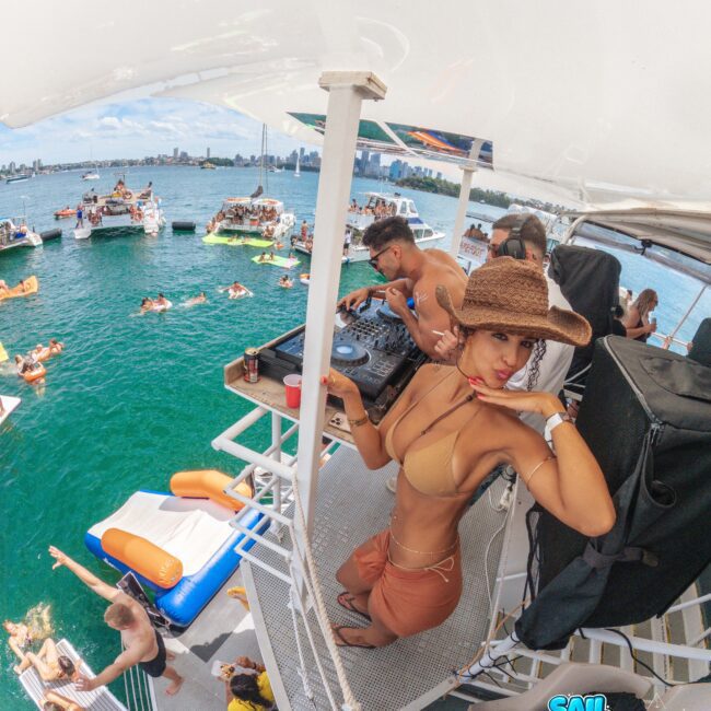 A woman in a brown bikini and sunhat poses on a boat deck near a DJ, overlooking a lively party with people swimming and relaxing on floats in the water, with city buildings in the background.
