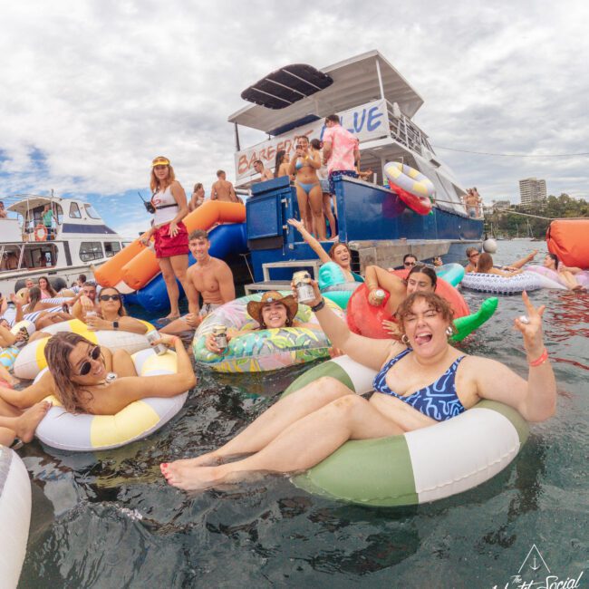 A group of young people in colorful swimwear relax on inflatable pool floats in the water, smiling and posing for the camera near a party boat with "BAY OF BLUE" written on it. The mood is festive and joyful.