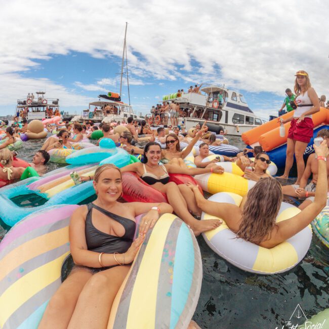 A large group of people relax on colorful inflatable pool floats in the water near boats under a partly cloudy sky, enjoying a lively party atmosphere.