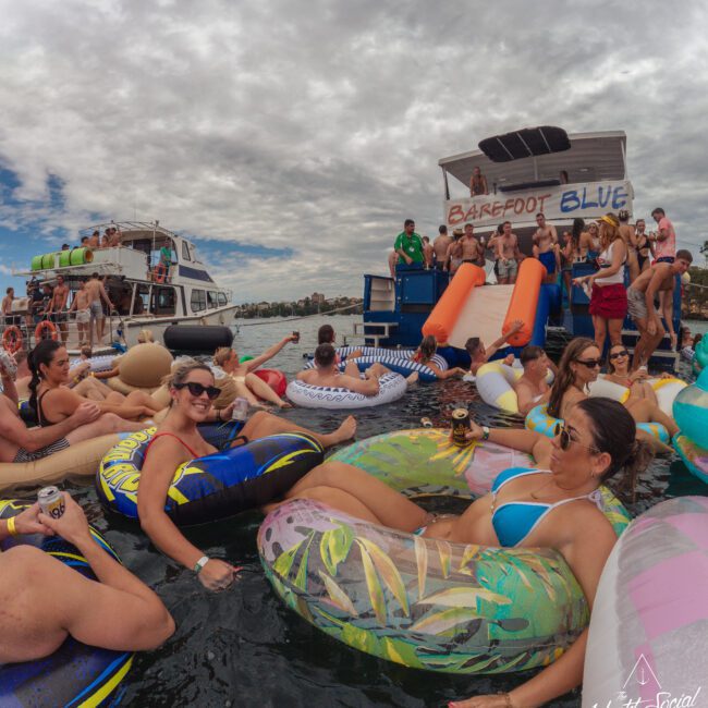 People in swimwear relax on colorful inflatables in the water near a party boat named "Barefoot Blue," with others socializing on the boat and another vessel in the background under a cloudy sky.