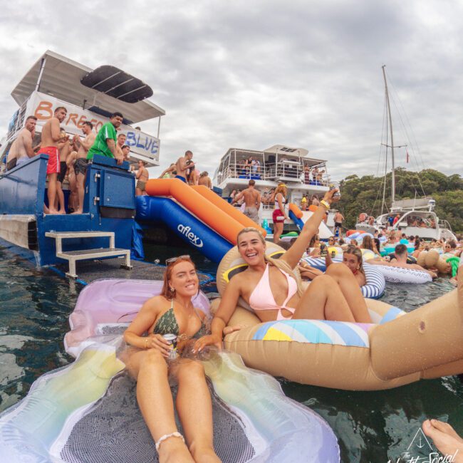 Two women relax on inflatable floats in the water near a crowded party boat, surrounded by people and more inflatables, with boats in the background under a cloudy sky.