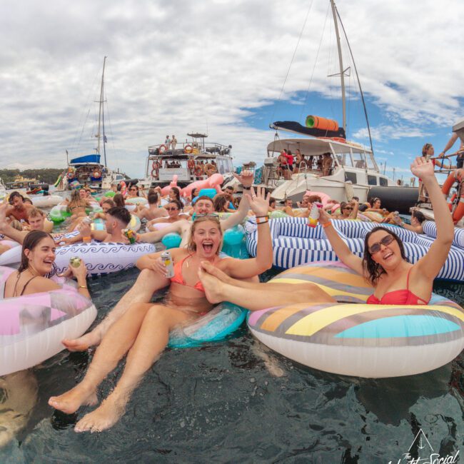 A group of people relax and have fun on colorful inflatable floaties in the water near docked yachts, laughing, smiling, and raising their hands during a lively outdoor party under a partly cloudy sky.