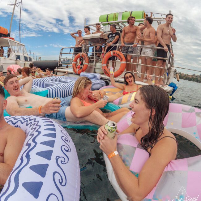 A group of people relax on colorful inflatables in the water near a boat, laughing, talking, and holding drinks. Others stand or sit on the boat, enjoying the sunny weather. The mood is festive and social.