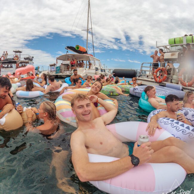 A group of people relax on colorful inflatable floaties in the water near anchored yachts, enjoying drinks and socializing under a partly cloudy sky. The atmosphere is lively and festive.