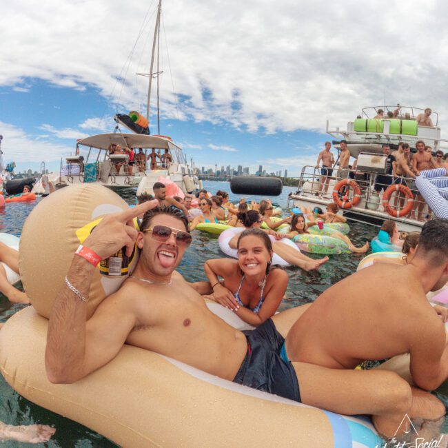 A shirtless man wearing sunglasses and holding a drink poses with a smiling woman on a large pool float in a crowded water party, with people on floats and boats in the background under a partly cloudy sky.