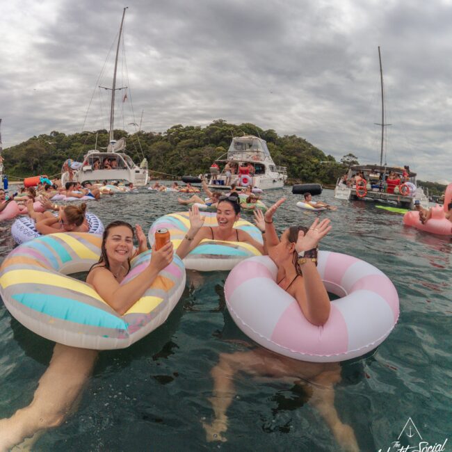 Three women smiling and waving while floating on colorful inflatable rings in the water, surrounded by other people and boats under a cloudy sky. A "Yacht Social Club" logo is visible in the corner.