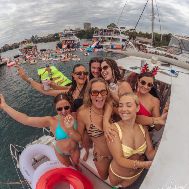 A group of smiling women in swimsuits pose together on a yacht, holding drinks and wearing sunglasses, with a festive, crowded boat party and cityscape in the background.