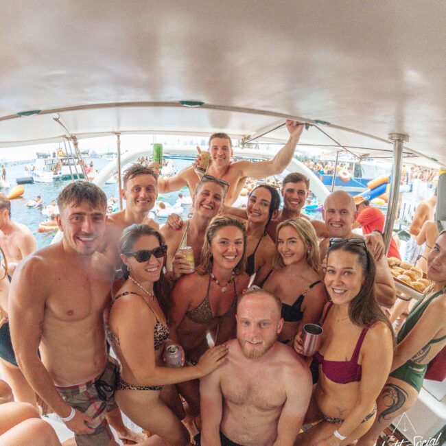 A group of people in swimwear smile and pose together on a boat deck during a sunny day, surrounded by water, other boats, and inflatable floats in the background.
