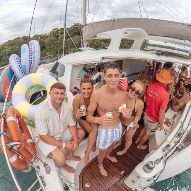 A group of young adults in swimwear relax and smile on a yacht deck, holding drinks and snacks. Inflatable pool floats and life rings are visible, with more people socializing in the background near the water.
