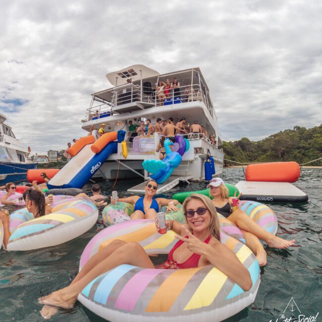 A group of people relax on inflatable pool floats in the water near a large party boat. Some people are on the boat, others enjoy the water, and everyone appears to be having fun under a cloudy sky.