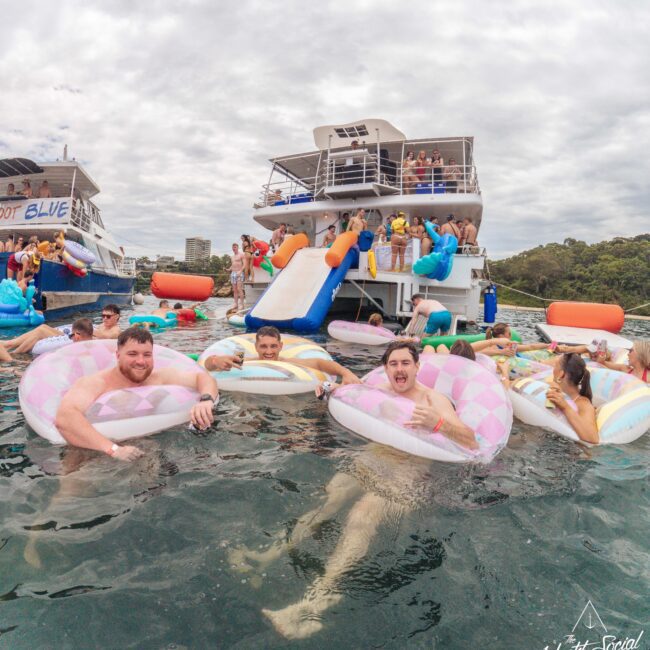 A group of people relax on inflatable pool floats in the water near two party boats, with others mingling, swimming, and enjoying a lively atmosphere under a cloudy sky. "Yacht Social Club" logo is visible in the corner.